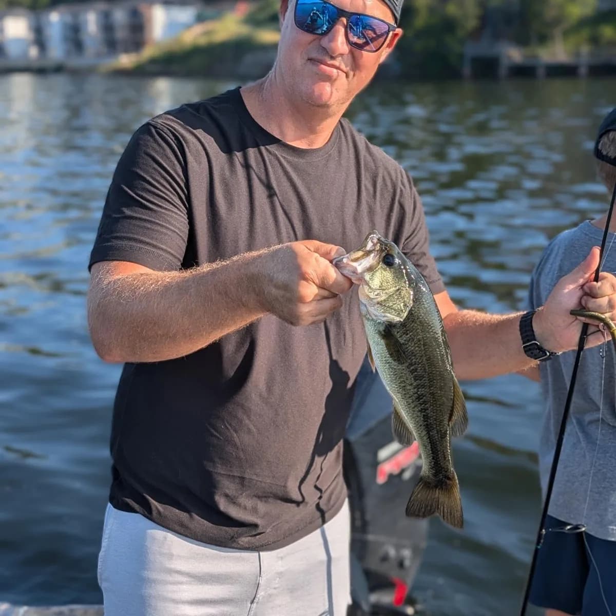 An angler holding a largemouth bass with a partner fishing behind