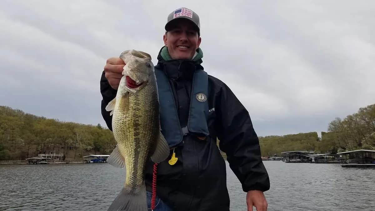 An angler in a dark jacket holding a largemouth bass on an overcast February day