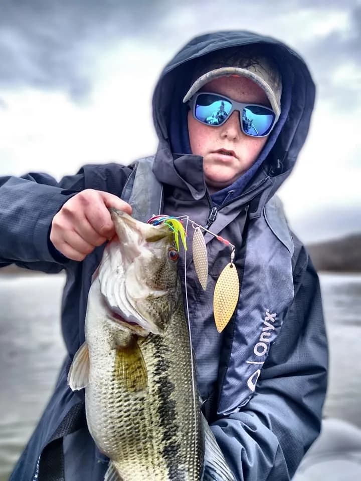 A young angler with his hood up holding a largemouth bass in the grey cold