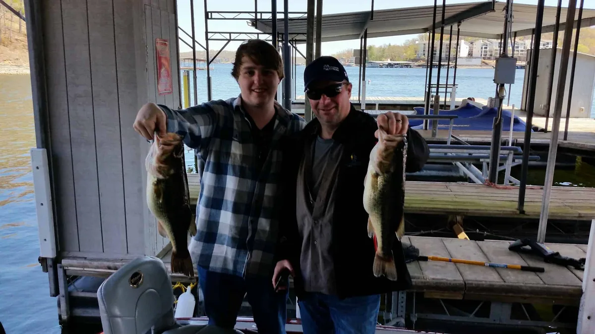 Two anglers in jackets each holding a largemouth bass under a boat dock