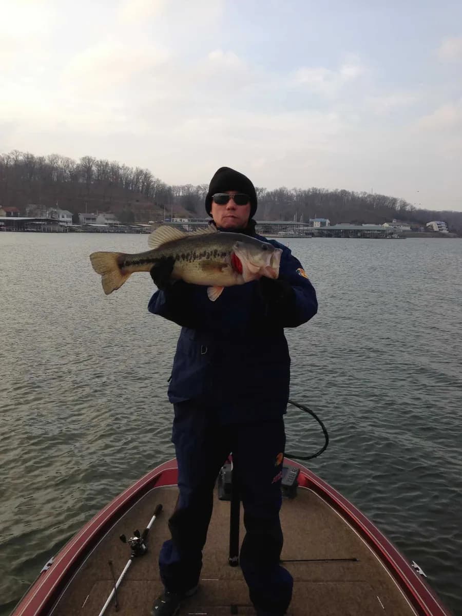 An angler in a heavy coat and beanie holding a largemouth bass on a bare-tree January day