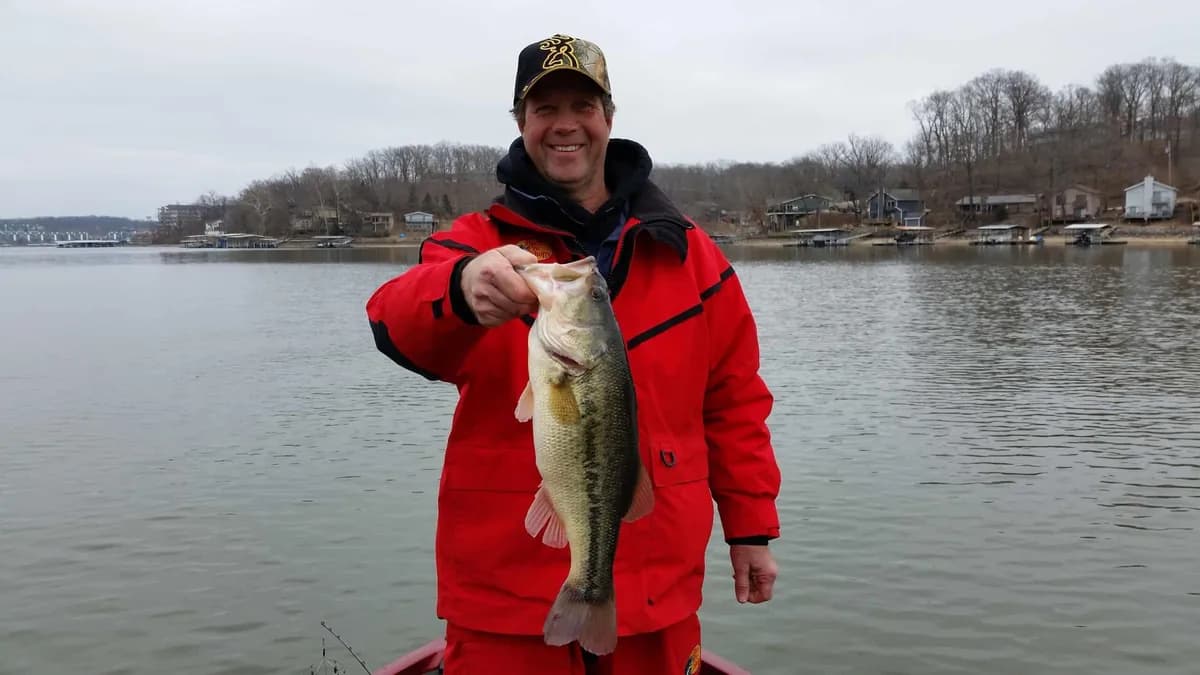 An angler in a red winter coat holding a heavy largemouth bass against a bare shoreline