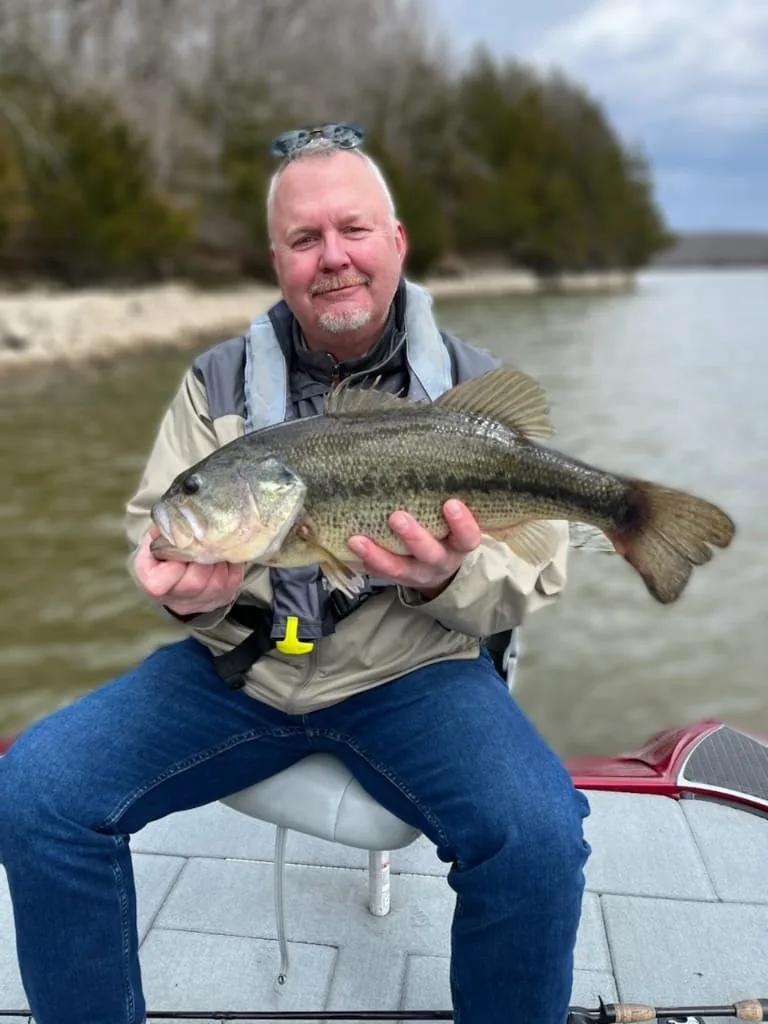 An angler seated on the deck with a largemouth bass, rocky bare shoreline behind