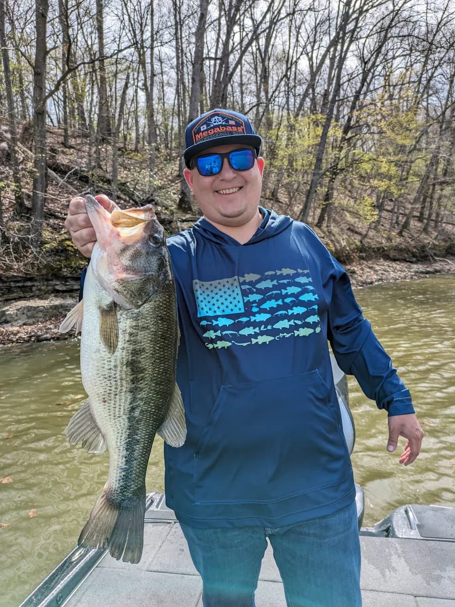 An angler in a camo hoodie holding a largemouth bass among bare March trees