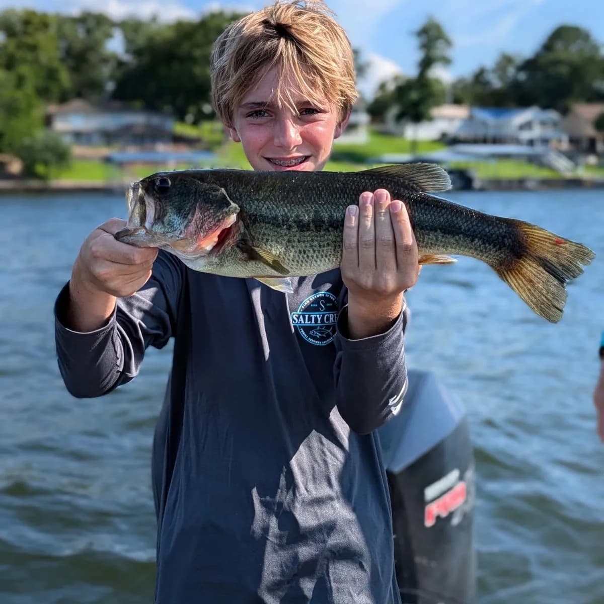 A young angler holding a largemouth bass with both hands, houses along the far shore