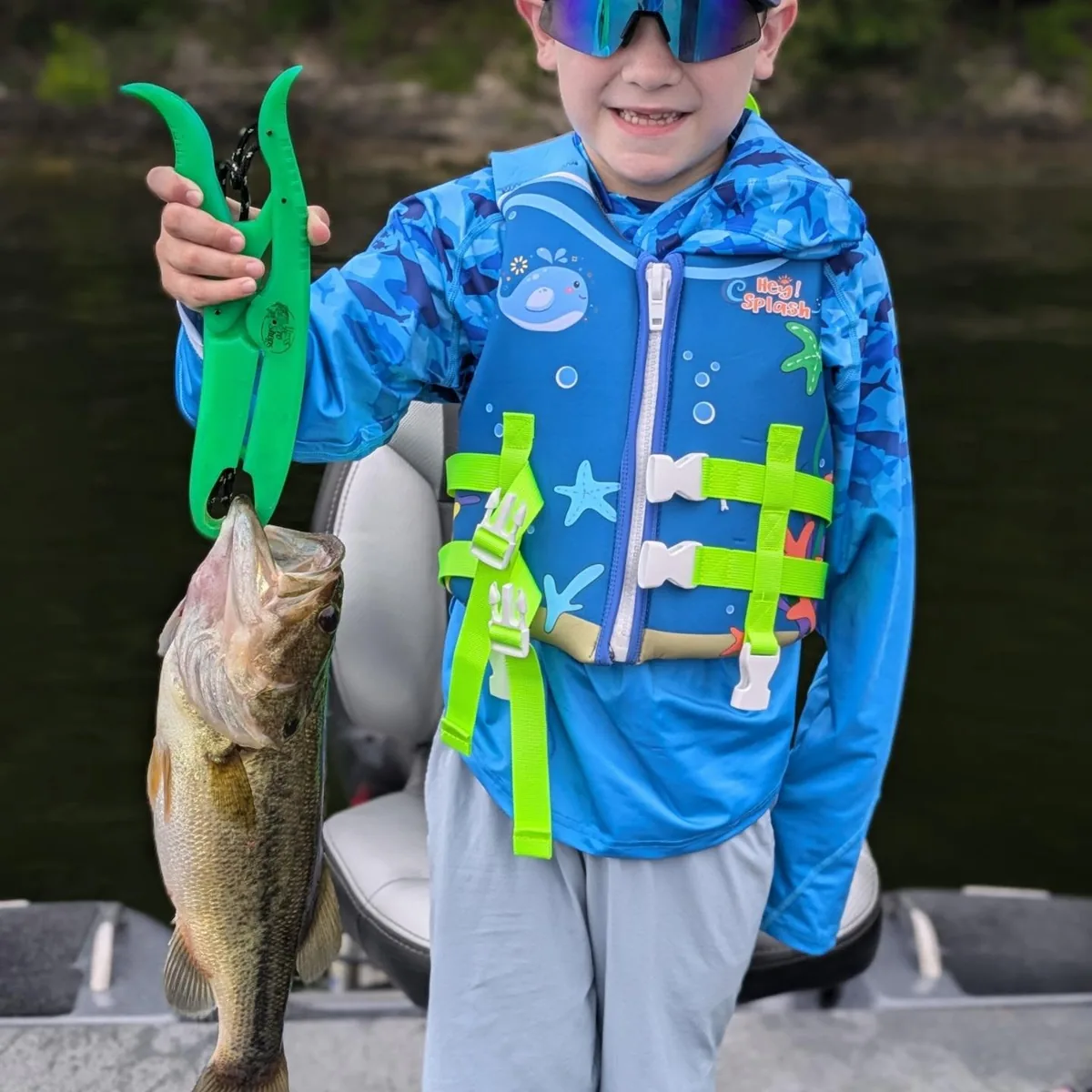 A young angler in a life jacket lifting a largemouth bass on a fish gripper