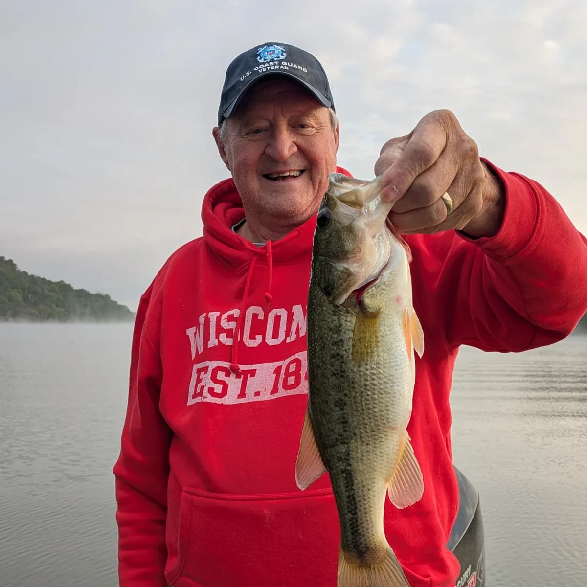 An angler in a red hooded sweatshirt holding a largemouth bass over misty water