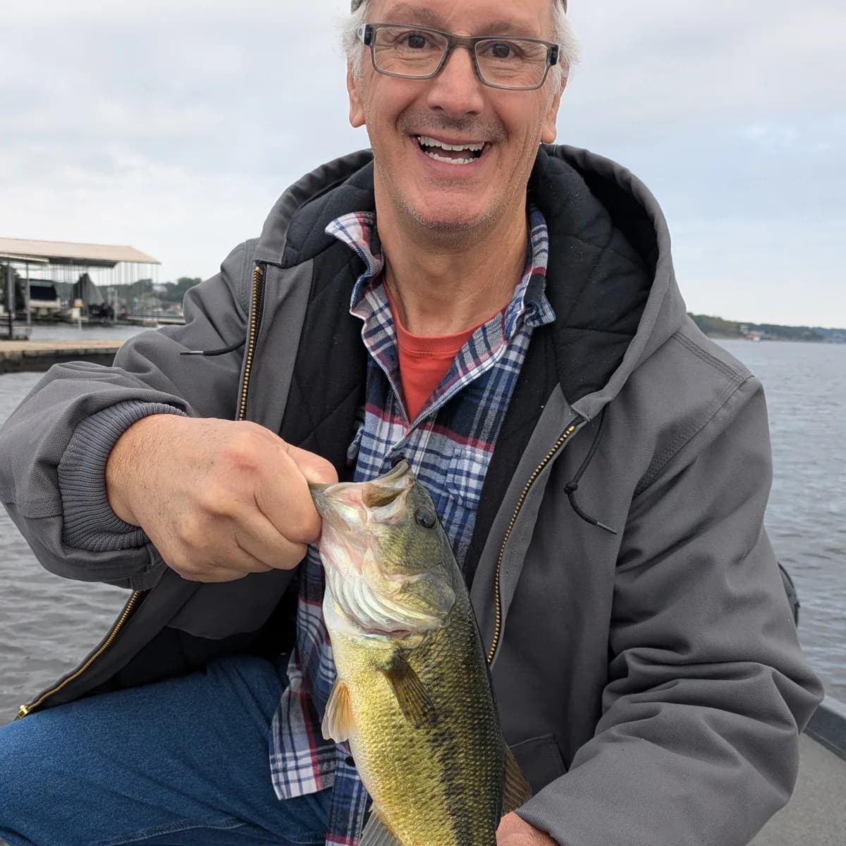 An angler kneeling on the deck with a largemouth bass, boat docks on the far bank