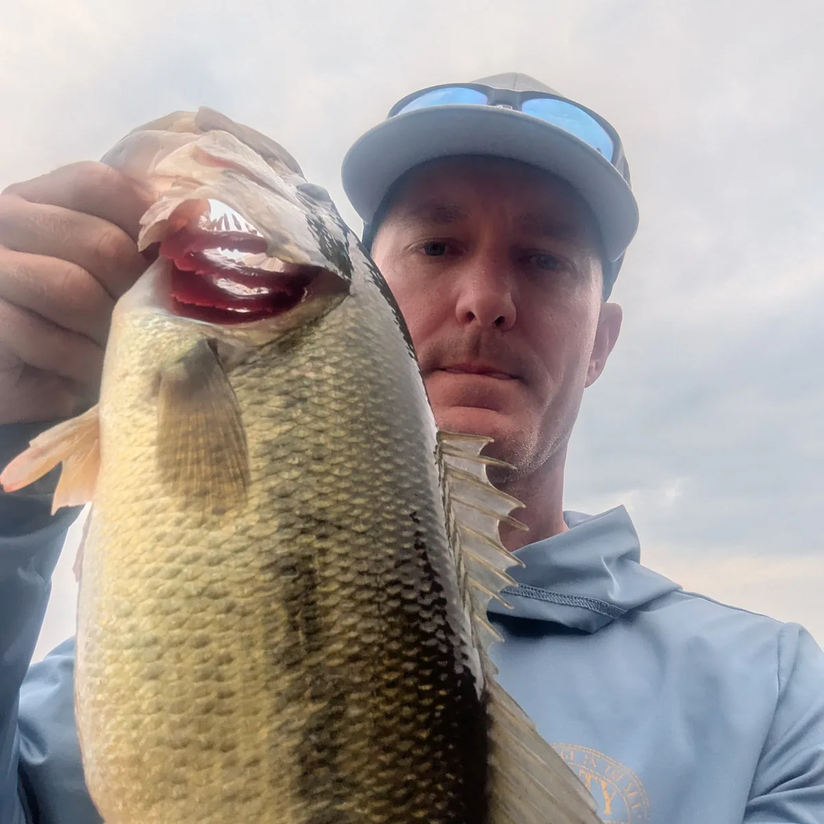 An angler in a sun shirt and cap holding a largemouth bass up to the camera