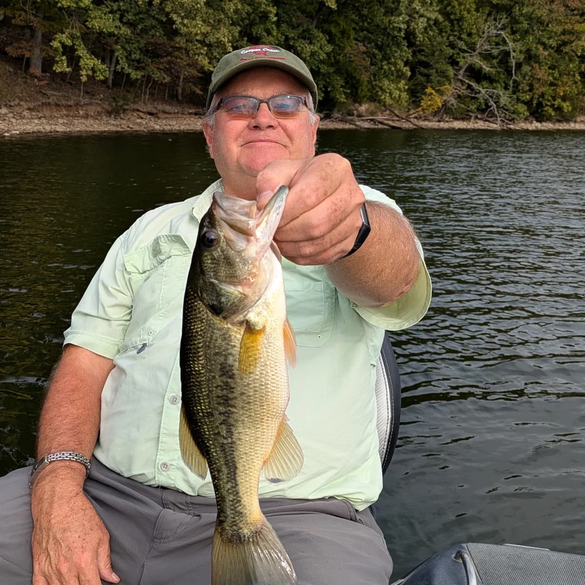 An angler seated on the boat holding out a largemouth bass, autumn timber behind